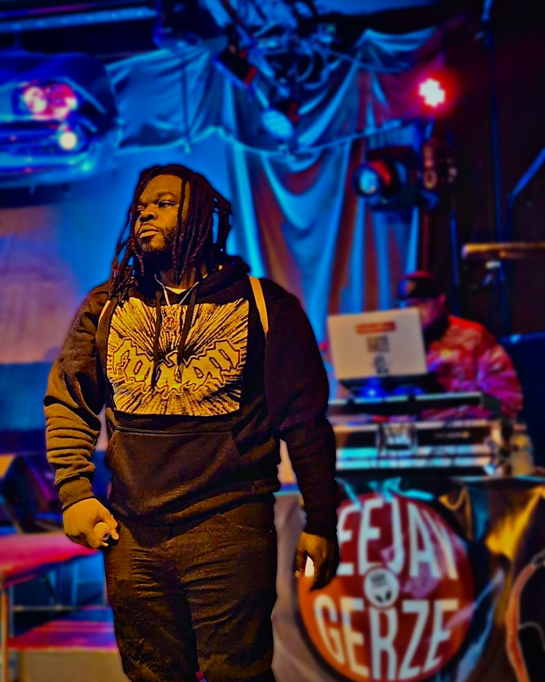 Person with dreadlocks wearing a hoodie on stage with DJ in background, colorful stage lighting.