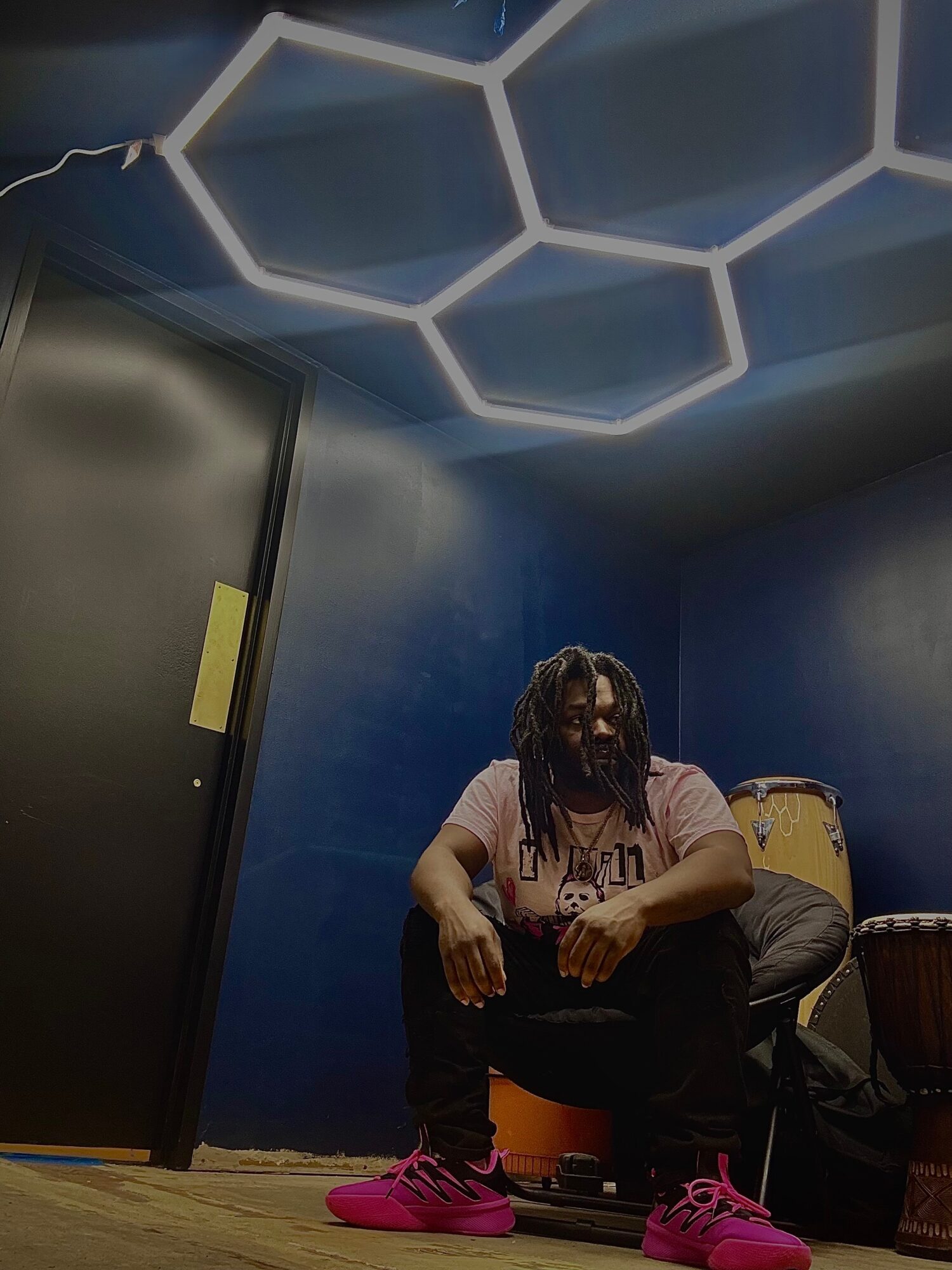 Person with dreadlocks sitting on a chair in a room with blue walls and hexagon-shaped ceiling lights.