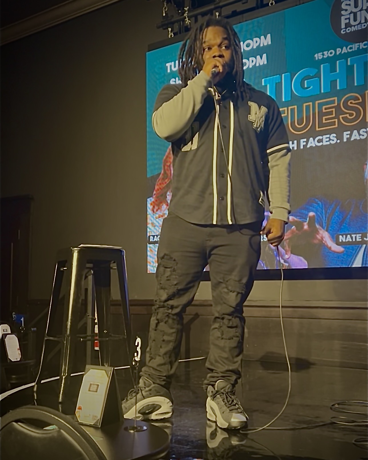 Person with long hair and glasses holding a microphone on stage, with a large screen behind displaying text and images.