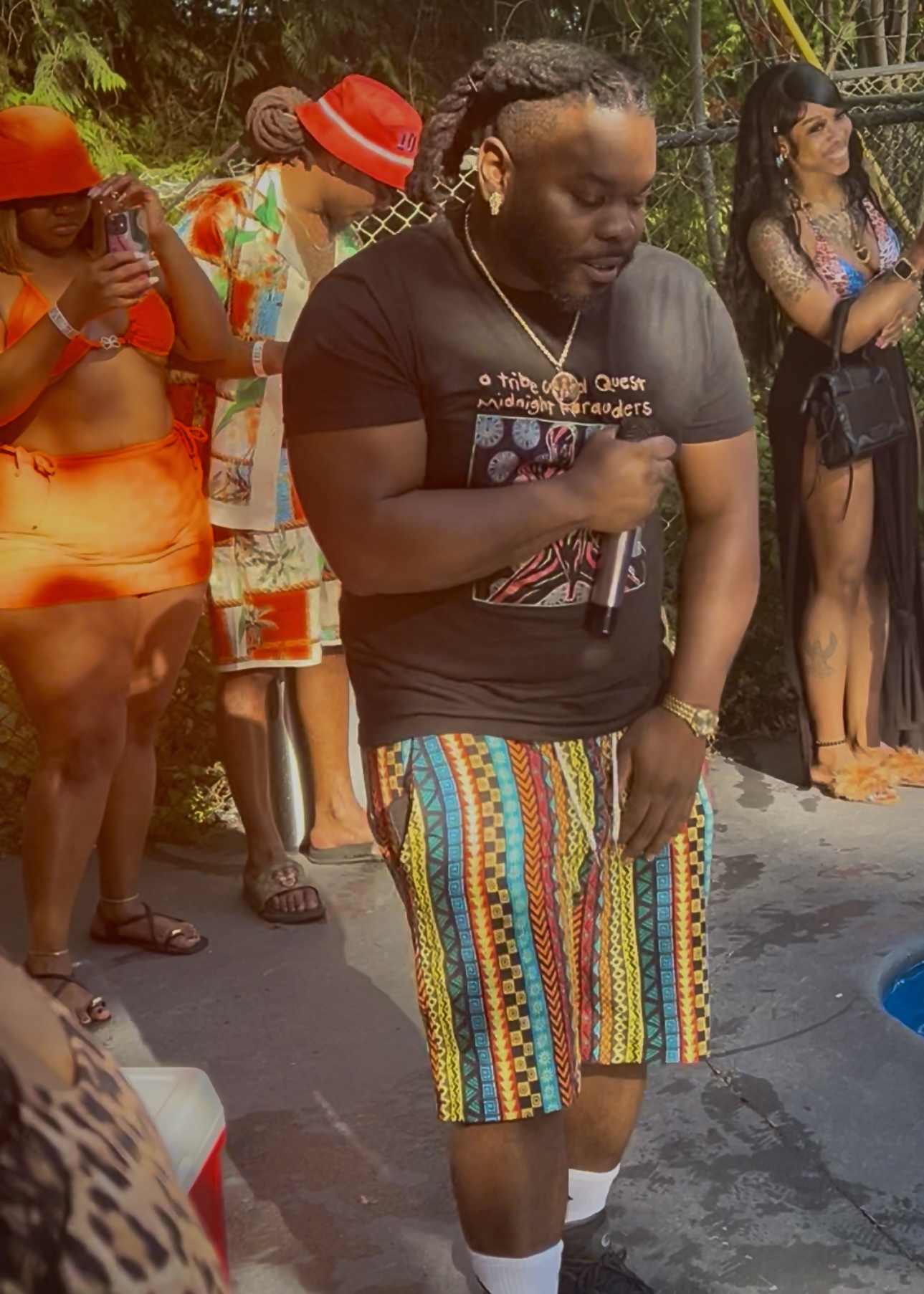 Man with braided hair wearing a black t-shirt and colorful striped shorts, holding a microphone outdoors.