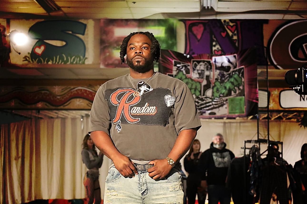 Man with beard and long hair standing in front of colorful graffiti art, wearing a gray t-shirt and jeans.