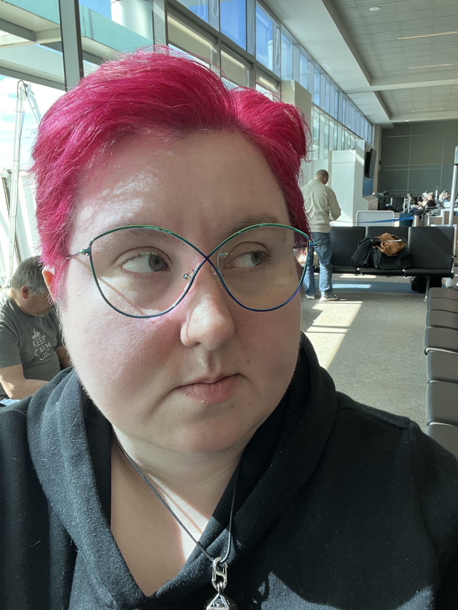 Person with bright pink hair and glasses in an airport terminal, looking to the side, with large windows and seating area behind.