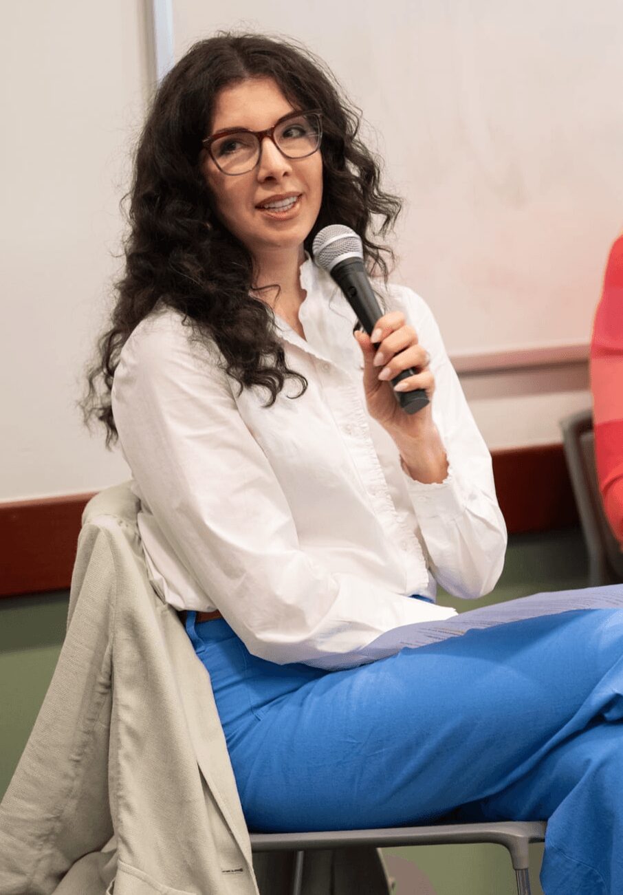 Woman with glasses holding a microphone, sitting on a chair, wearing a white shirt and blue pants, in a room with a whiteboard.