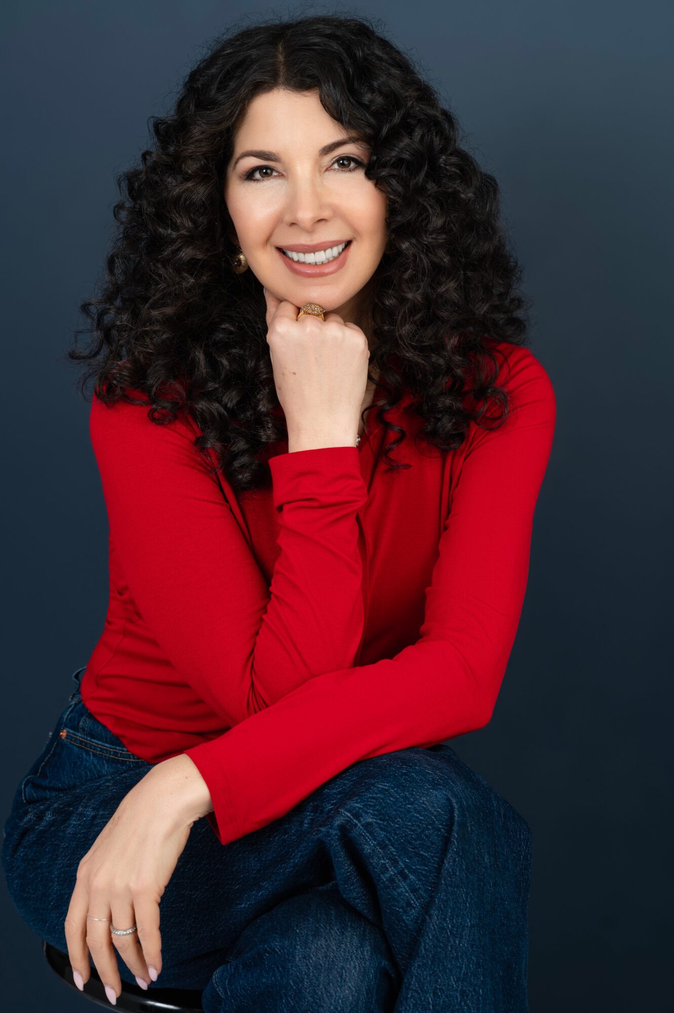Woman with curly hair smiling, wearing a red top, sitting with her chin resting on her hand, against a dark background.