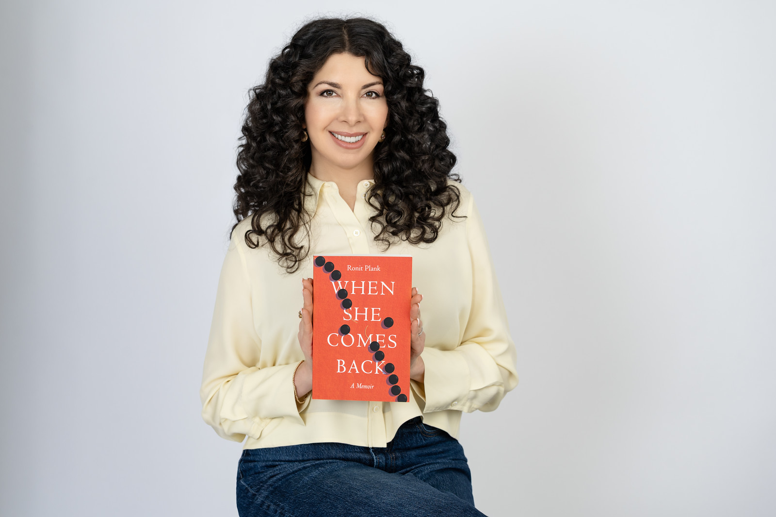 Woman with curly hair holding a red book titled 'When She Comes Back' smiling, wearing a cream blouse and jeans, against a plain background.