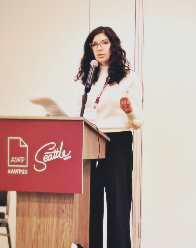 Woman with dark curly hair and glasses speaking at a podium with a microphone, wearing a white blouse and black pants.