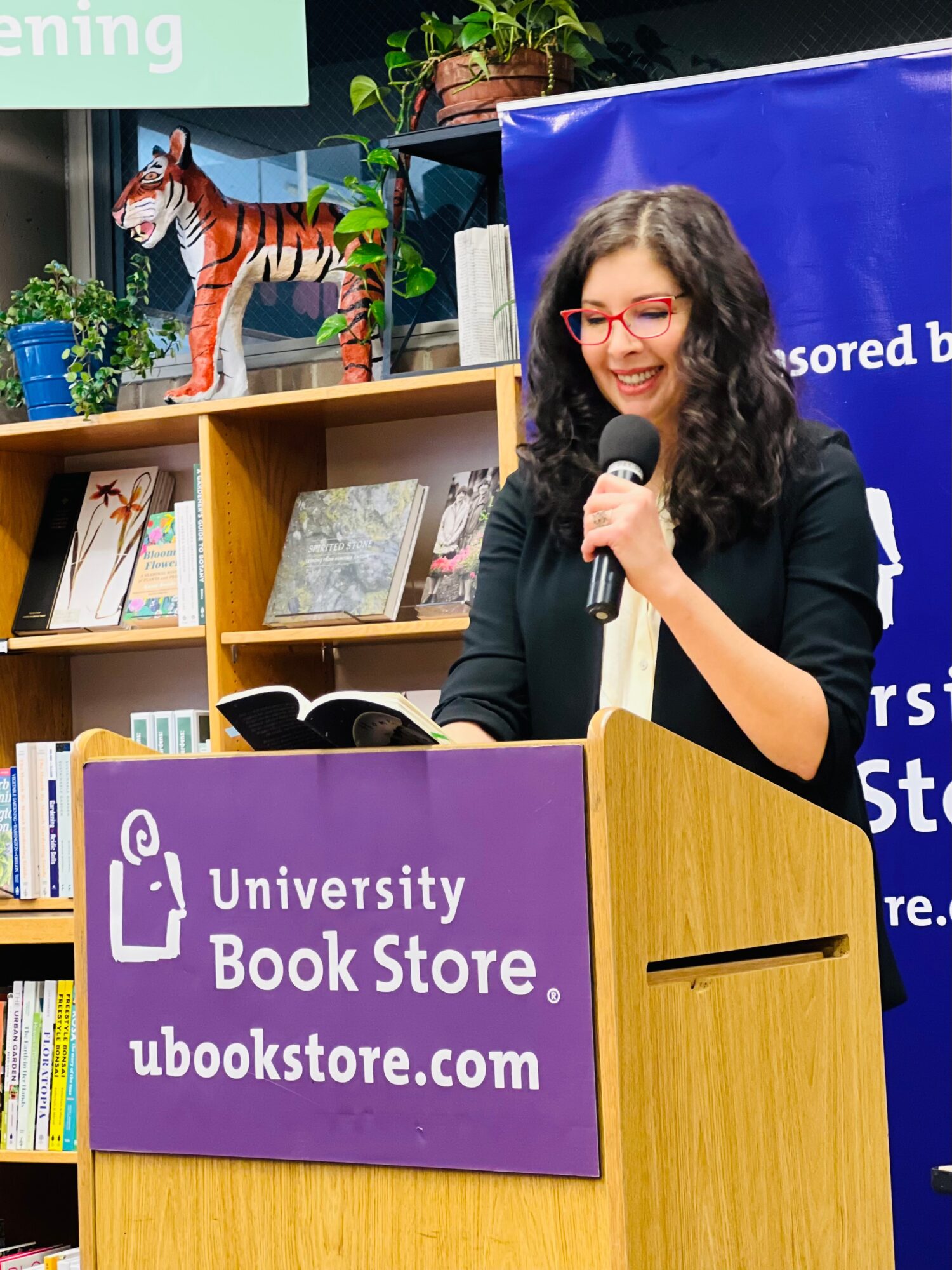 Woman with glasses speaking at a podium with a purple sign for University Book Store, books and decorations behind her.