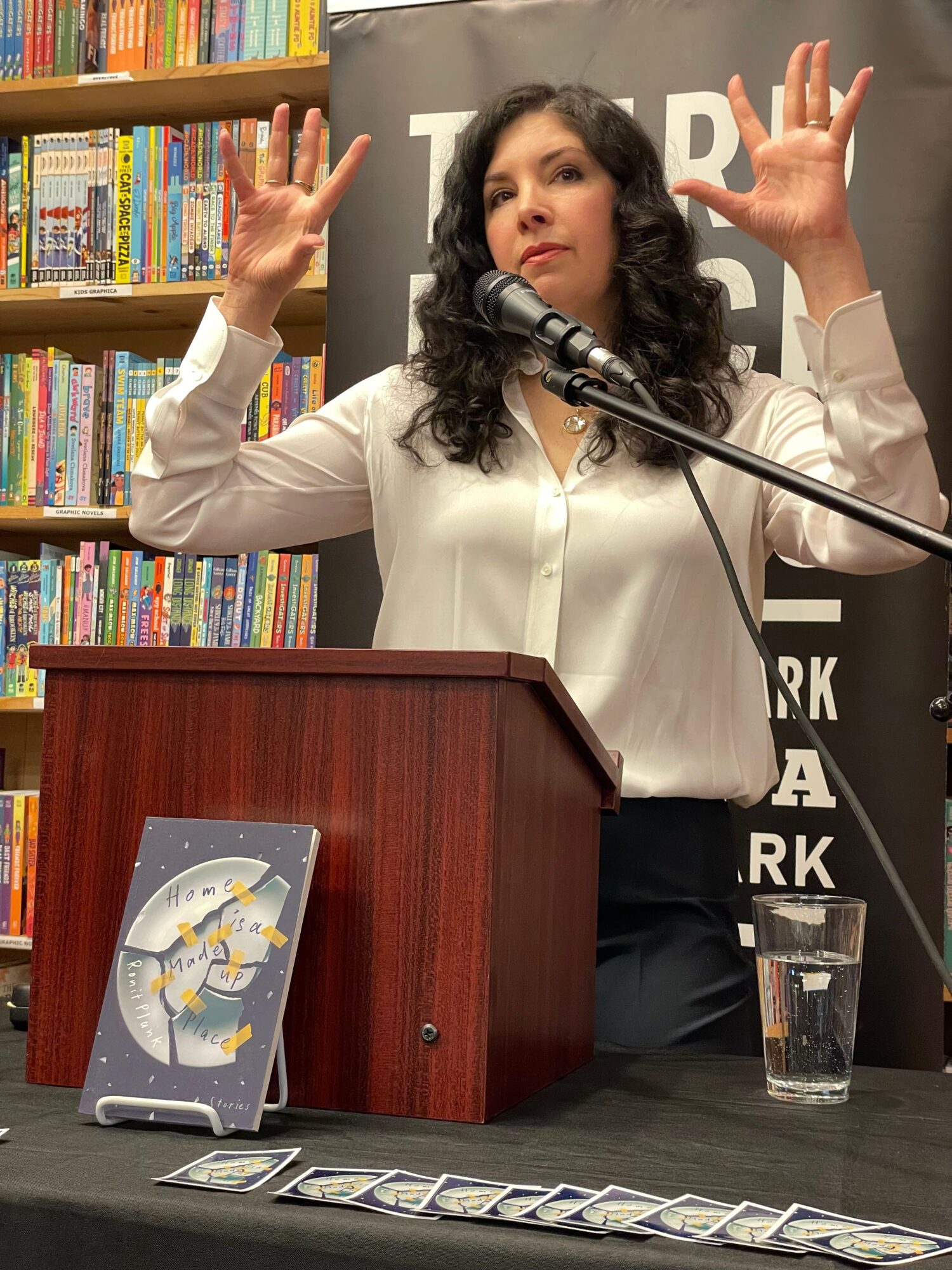 Woman speaking at a podium with bookshelves in the background, gesturing with her hands, and a book on the table.