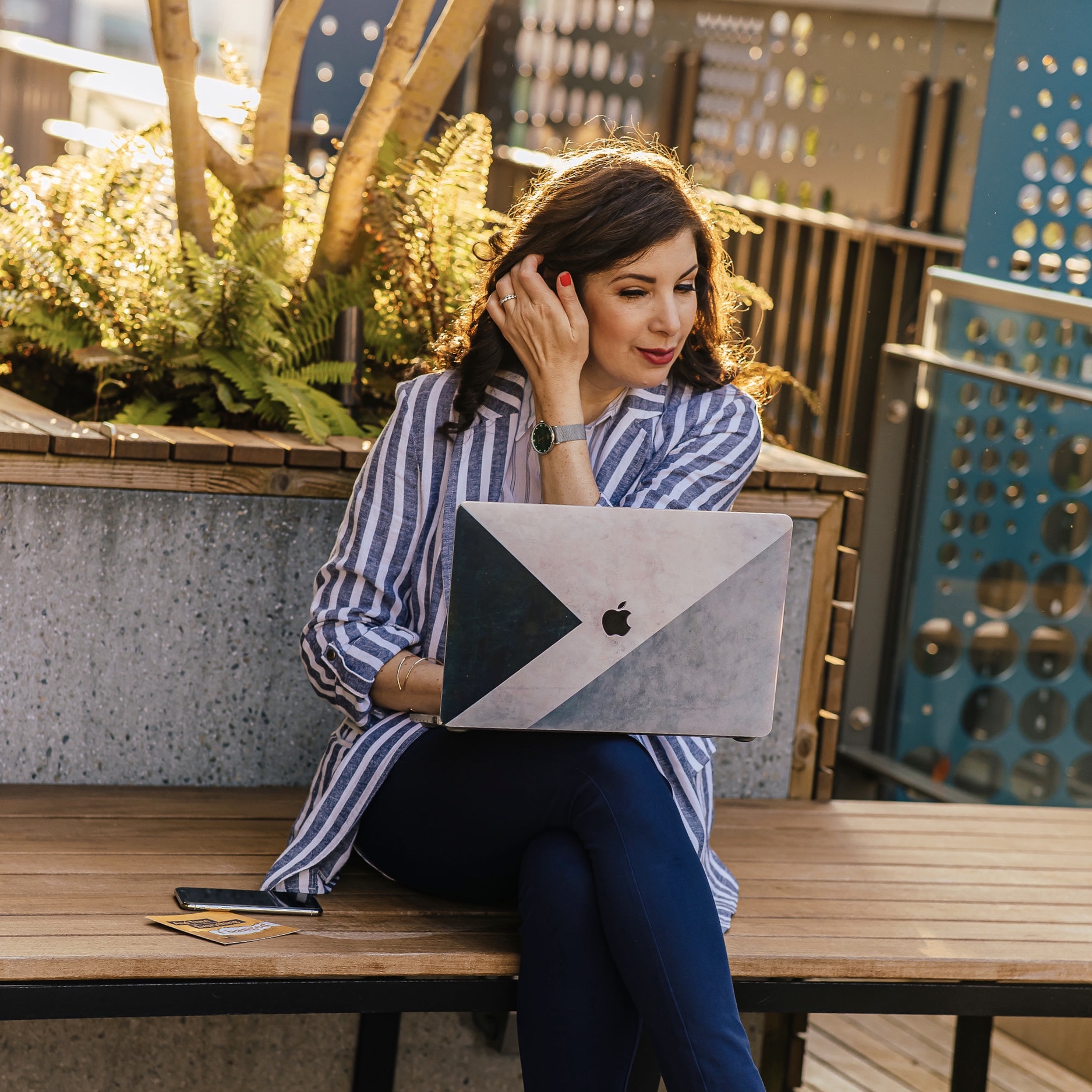 Woman sitting on a wooden bench using a laptop outdoors, with plants and a fence in the background.