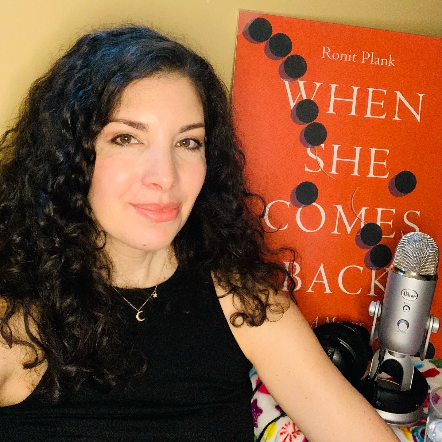 Woman with curly hair taking a selfie next to a book and microphone, smiling at the camera.