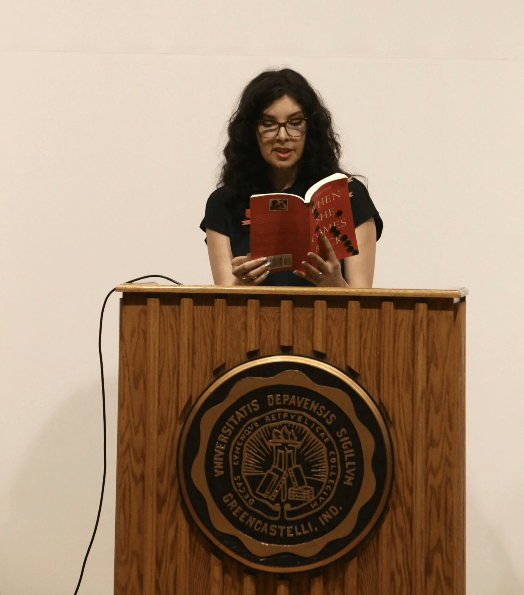 Woman with glasses reading a book at a wooden podium with university emblem, against a plain white wall.