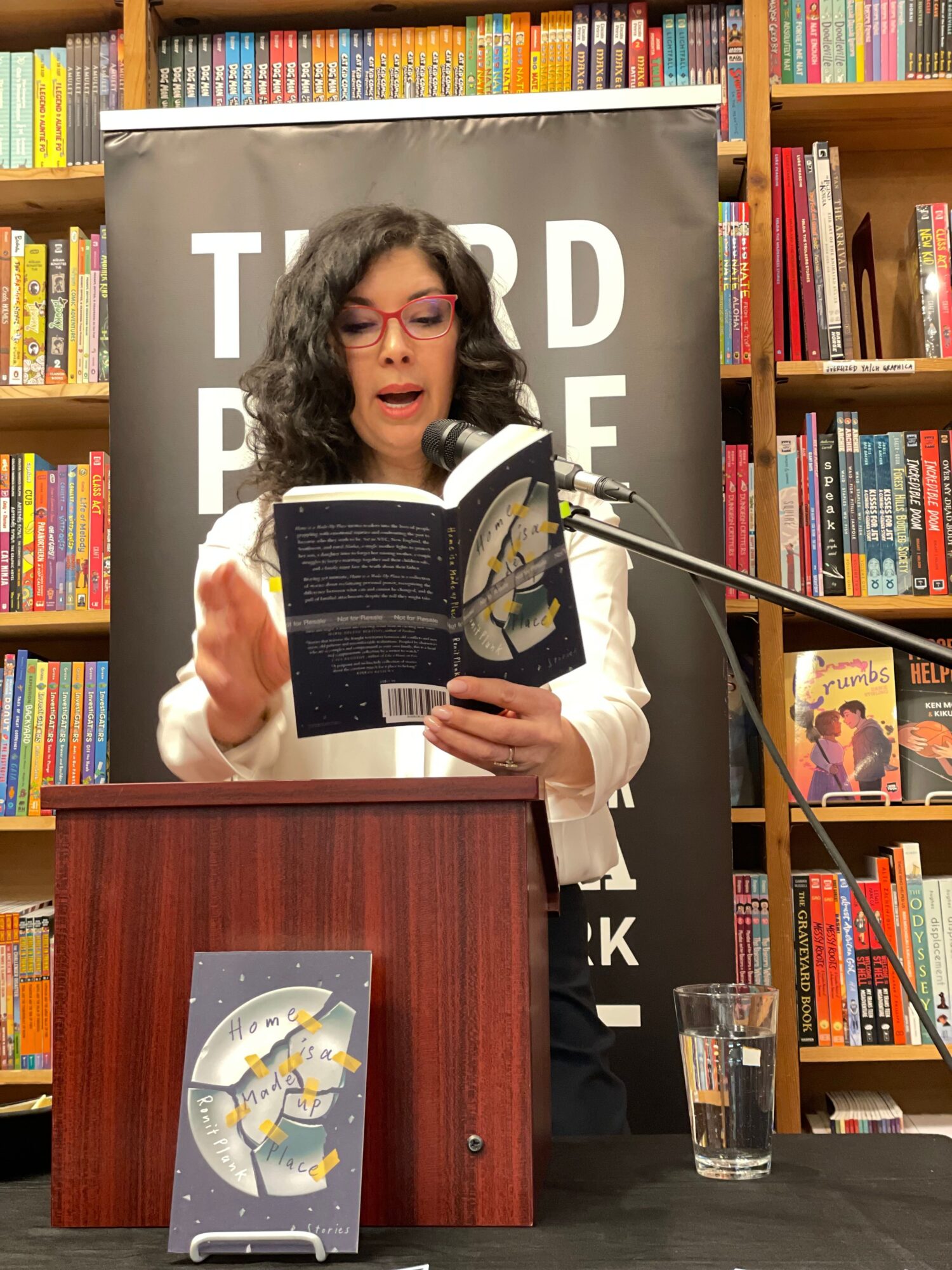 Woman speaking at a podium in a bookstore, holding a book, with bookshelves in the background.