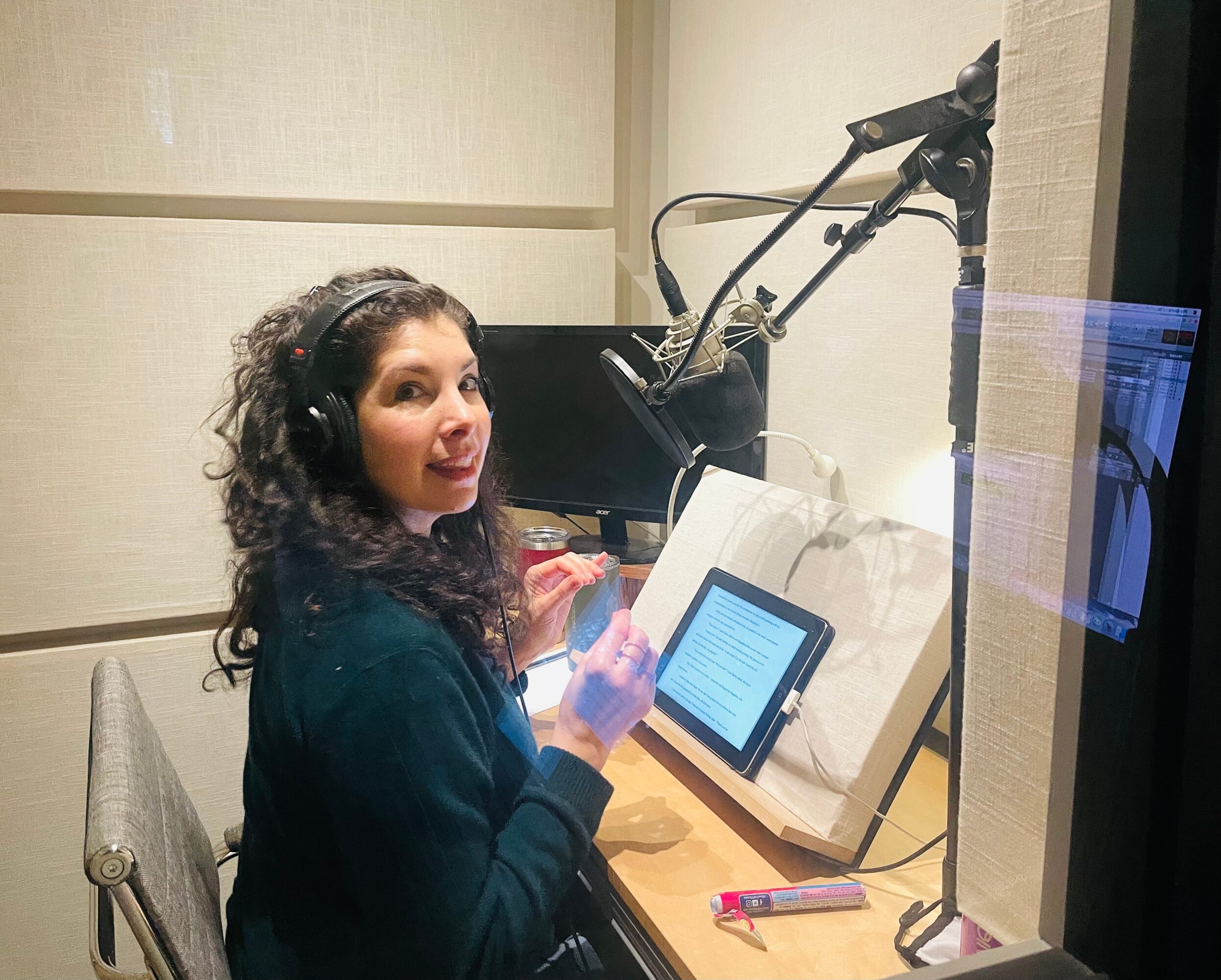 Woman with curly hair wearing headphones, sitting at a desk with a tablet and microphone in a recording studio.