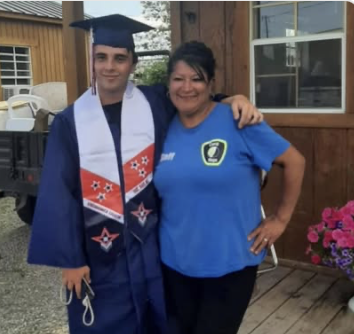 A young man in a graduation gown and cap stands next to a woman in a blue shirt, both smiling outdoors.
