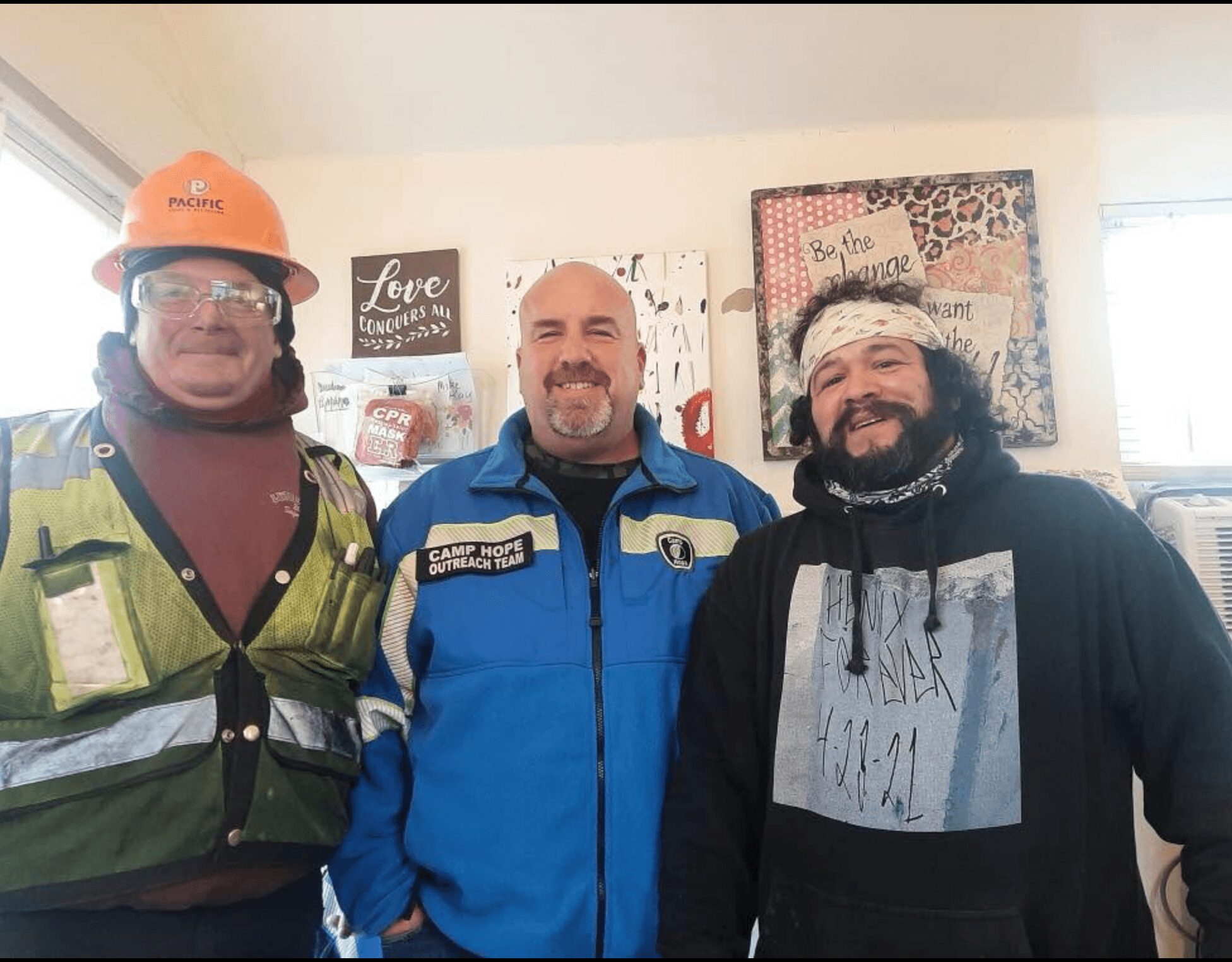 Three men standing indoors, smiling, with artwork and decorations on the wall behind them.