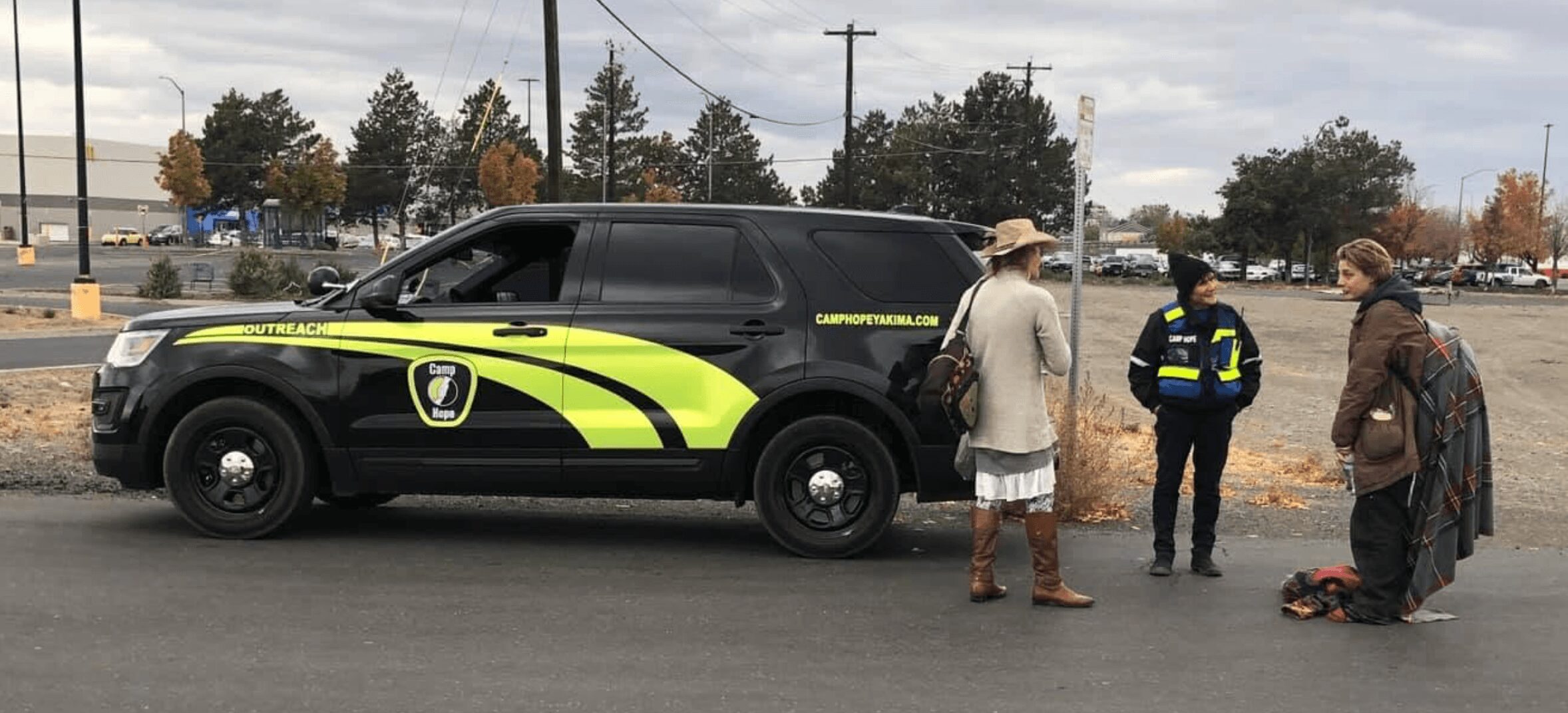 Four people standing near a black and yellow police vehicle on a street, with trees and buildings in the background.