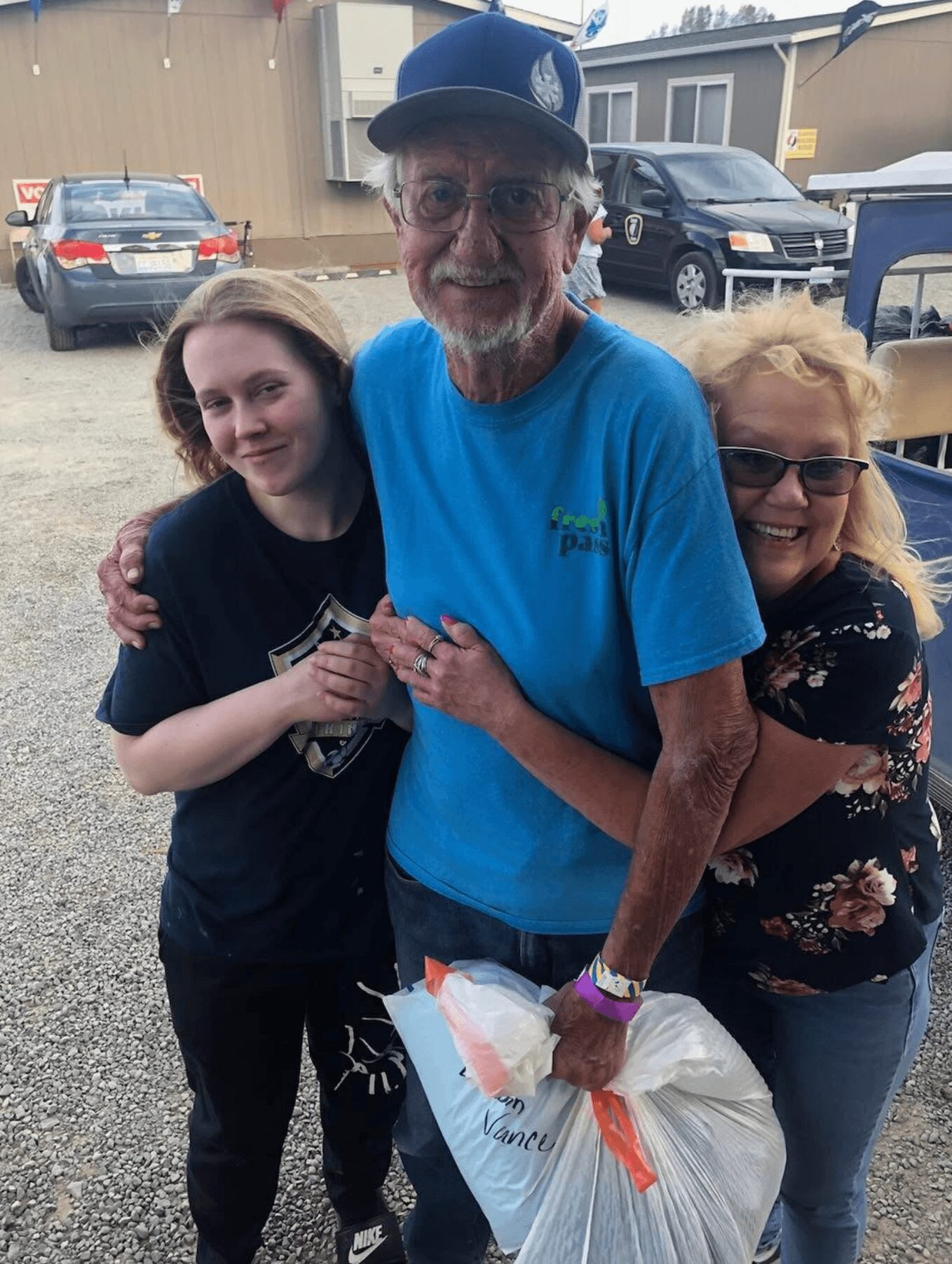 Three people smiling and hugging outdoors, with cars and buildings in the background.