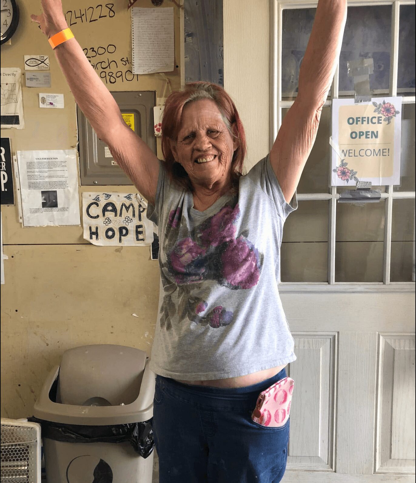 Woman with arms raised smiling, wearing a gray t-shirt with a colorful design, standing indoors near a door and window.