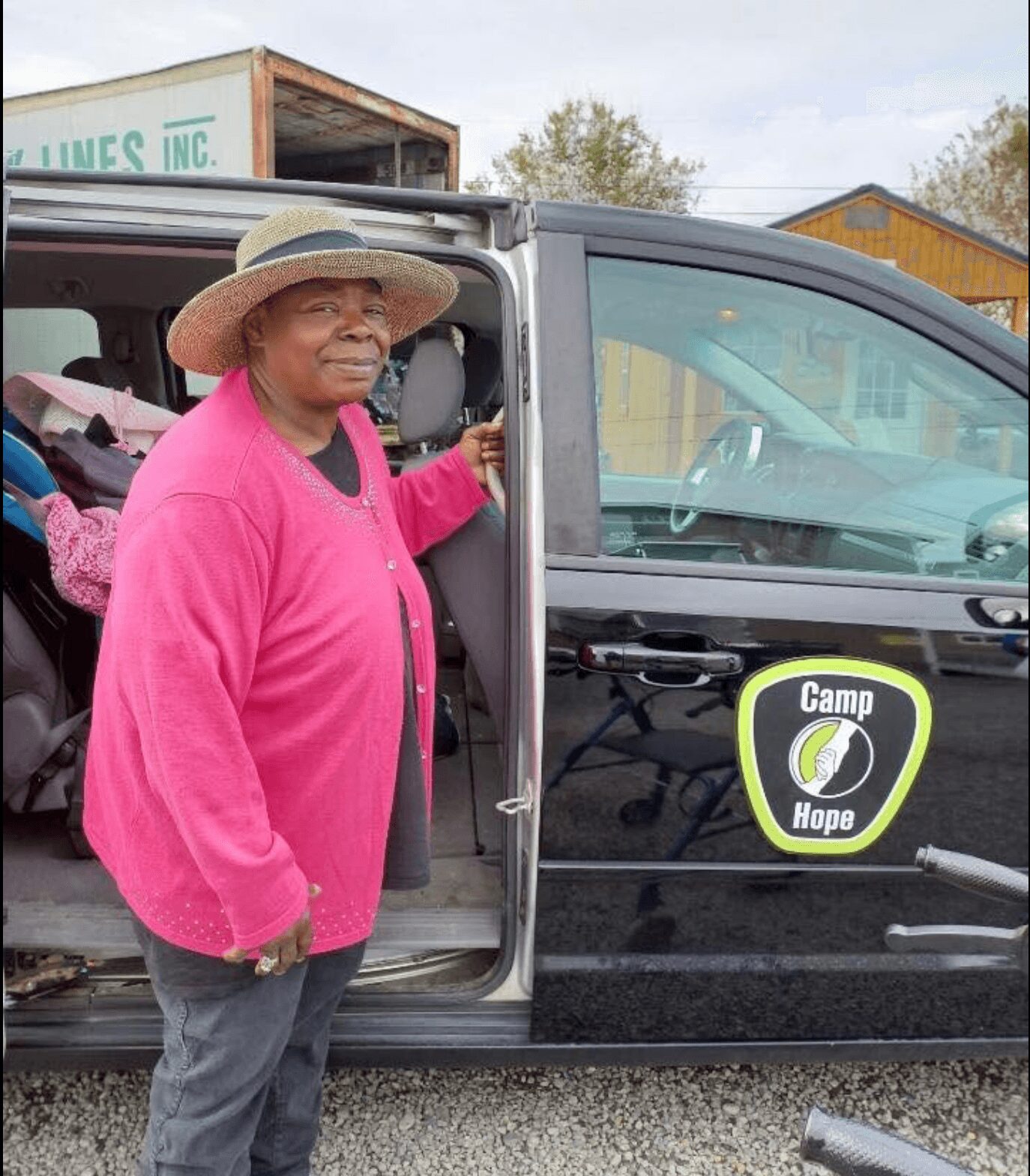 Older woman wearing a wide-brimmed hat and pink cardigan standing next to a vehicle with a 'Camp Hope' sign.